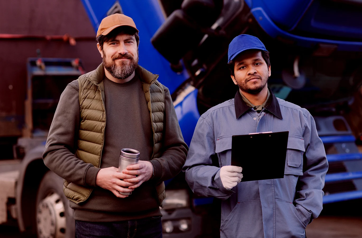 Two men stand outdoors near a large blue vehicle, one holding a coffee cup and the other holding a clipboard, wearing work attire.