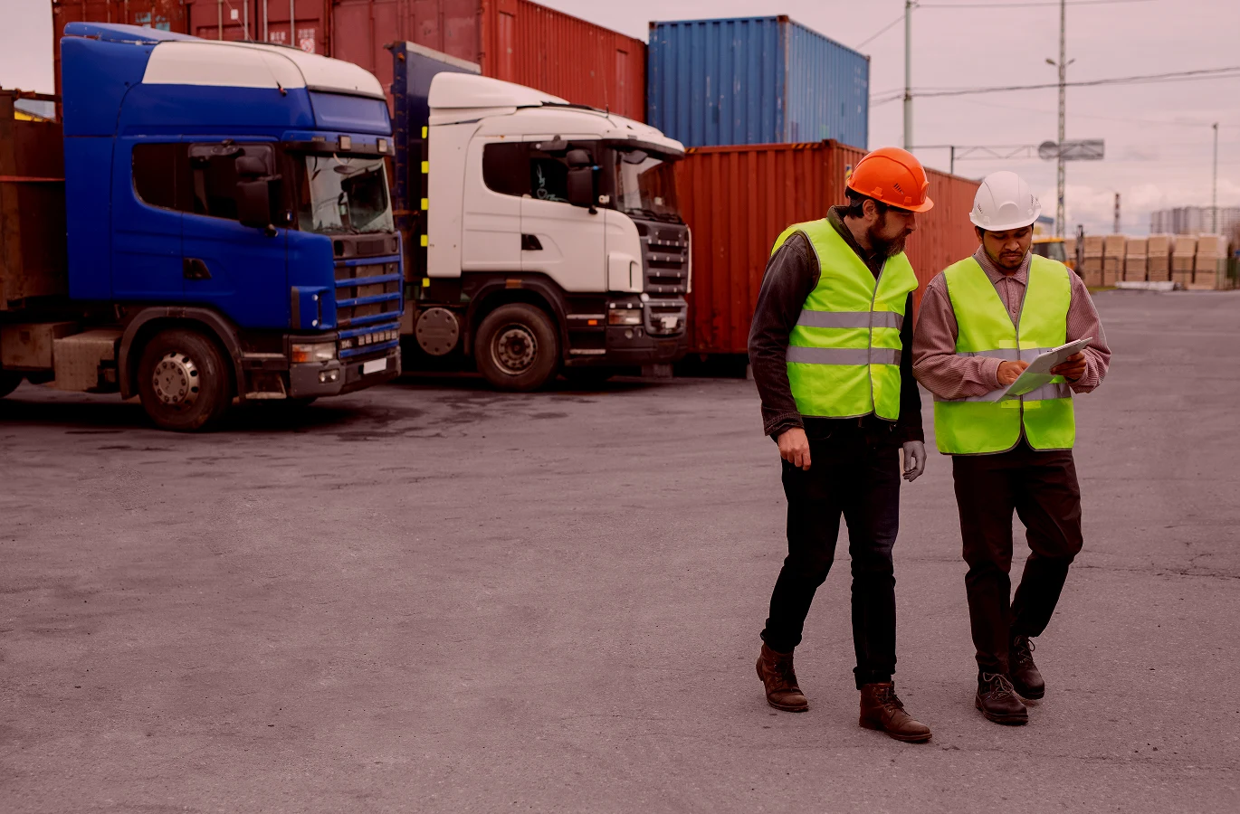 Two construction workers in safety vests and helmets discuss plans in a truck yard surrounded by containers and parked trucks.